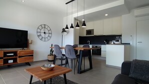 Ein modernes Wohnzimmer mit Flachbildfernseher, Holzmöbeln, einem Essbereich mit Stühlen und einer Küche mit weißen Schränken und einer schwarzen Arbeitsplatte.