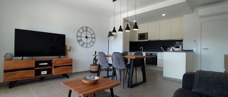 A modern living room with a flat-screen TV, wooden furniture, a dining area with chairs, and a kitchen with white cabinets and a black countertop.