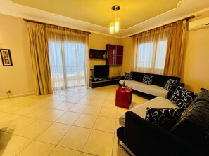 A spacious living room with a sofa, red ottoman, and a flat-screen TV.