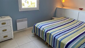 Un dormitorio con cama, cómoda, radiador, ventana y lámpara fijada en la pared.