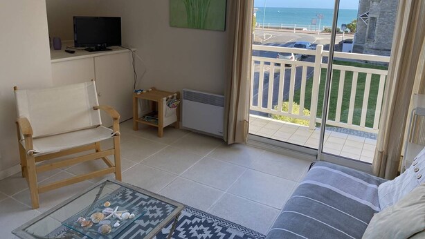 Sala de estar con televisor, una silla, una mesita con una revista y vista al mar a través de amplios ventanales.