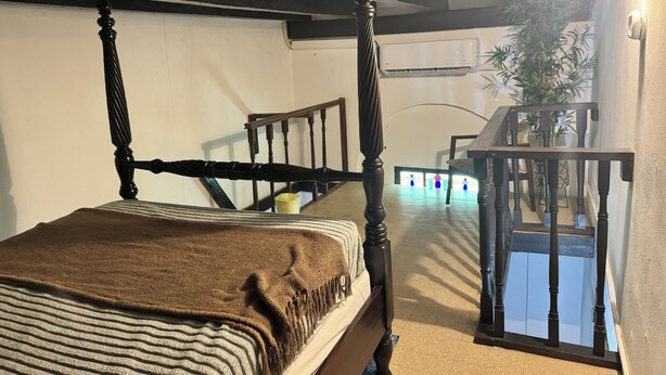 A bedroom with a four-poster bed, a small bedside table, and a nightstand. There is a window with blinds, a potted plant, and a staircase leading to an upper level.
