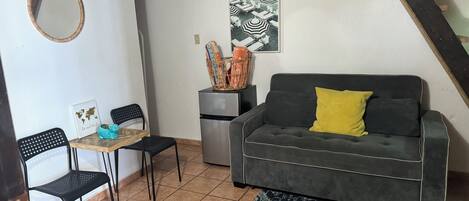 Ein kompakter Wohnbereich mit einer Couch, einem kleinen Tisch, zwei Stühlen, einem Kühlschrank und einem Wandbild.