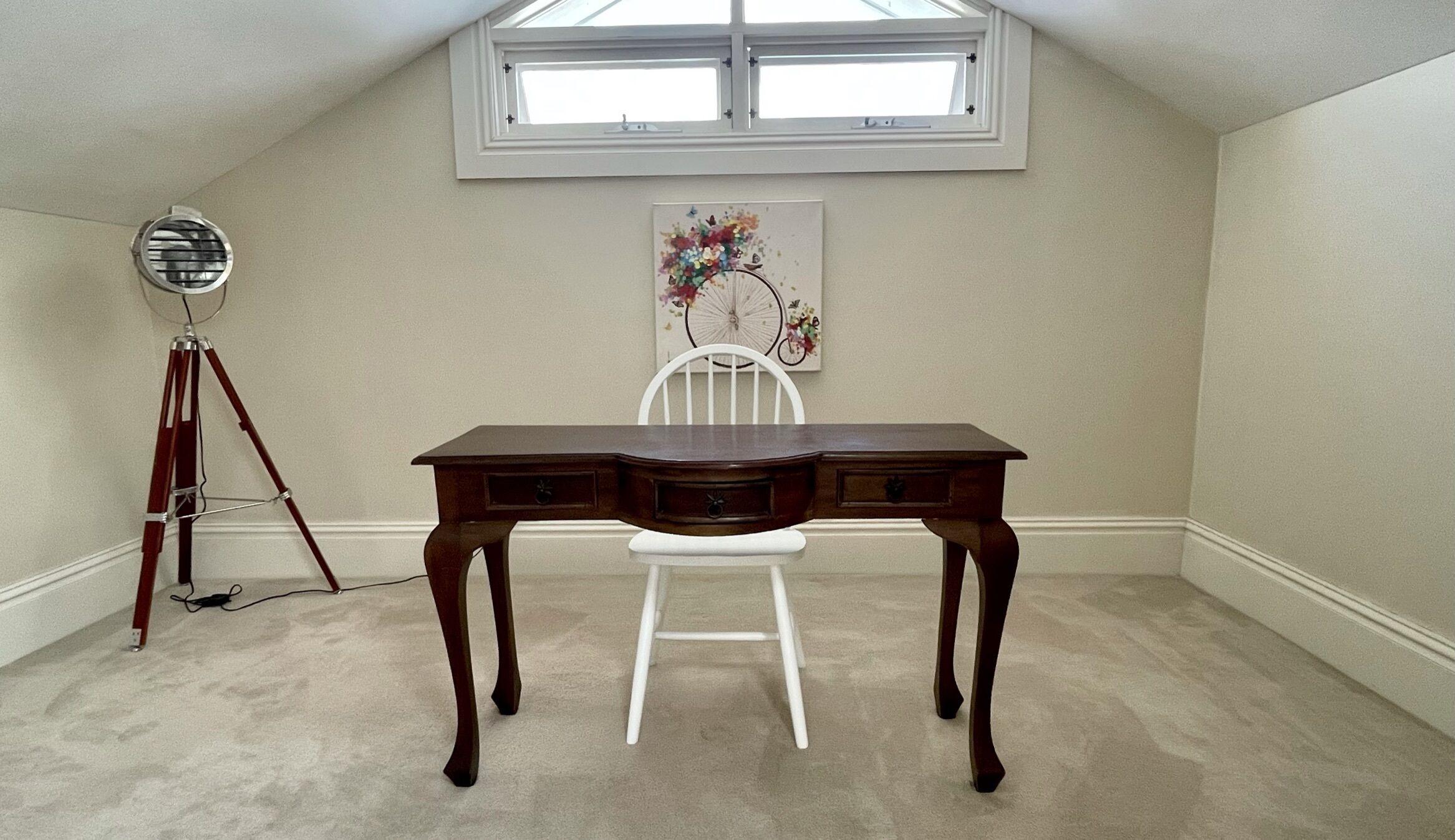 Una habitación con techo inclinado, un escritorio de madera con cajones, una silla blanca, una lámpara de pie con trípode y un ventanal.