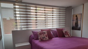 A bedroom with a bed, a window with blinds, a small white bench, and a painting on the wall.