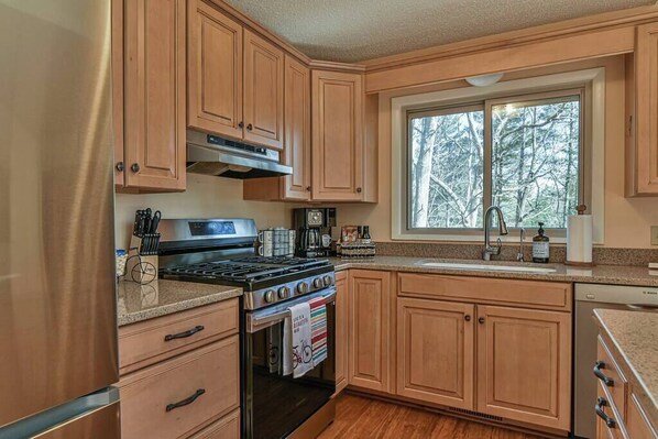 A kitchen with wooden cabinets, a stove, a sink, and a window with a view of trees.