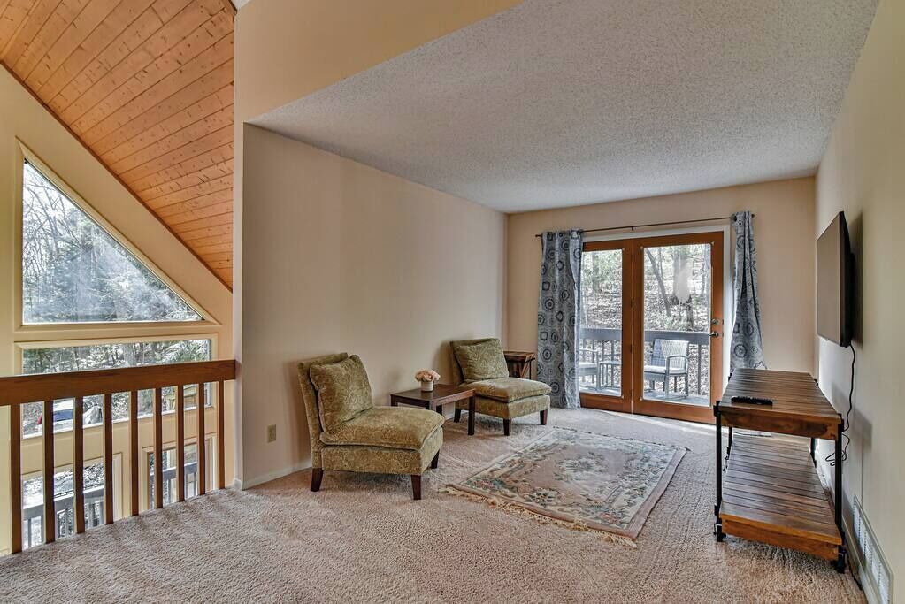 A room with a high ceiling, a large window, two armchairs, a small table, a rug, and a TV on the wall.