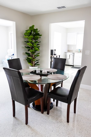 Un espace repas moderne avec une table en verre entourée de chaises noires, une plante en pot et une cuisine en arrière-plan.