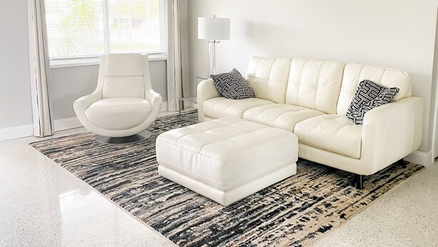 Una sala de estar moderna con un sofá de cuero blanco, un reposapiés a juego y un sillón blanco. Una alfombra estampada cubre el suelo, y se ve una ventana con persianas.