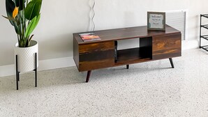 Una sala de estar moderna con un televisor de pantalla plana montado en la pared, un mueble de madera para entretenimiento con una revista, y una planta en maceta con hojas verdes.