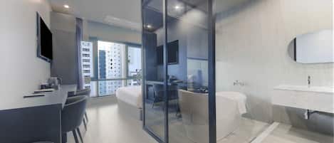 Une chambre d’hôtel moderne dotée d’une paroi en verre qui sépare l’espace de vie de la chambre à coucher. Elle comprend une télévision à écran plat, une table à manger avec des chaises, un canapé et une grande fenêtre offrant une vue sur la ville.