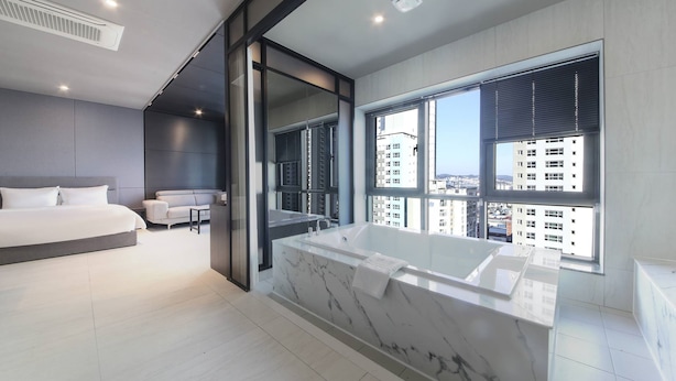 A modern bedroom with a large bed, a separate sitting area, and a bathroom with a large bathtub and city views. - Busan station city hotel (Busan)
