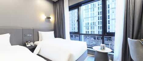A hotel room with two beds, a bedside table with a phone, a round table with a lamp, and a large window with a city view.