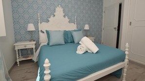 En pænt redt seng med hvid gavl og turkis sengetøj, et hvidt natbord med lampe, og tapetbaggrund med mønster.