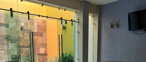 Un espacio interior moderno con una mesa y sillas de madera, una puerta de cristal que da al exterior, y un televisor montado en la pared.