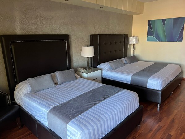 A hotel room with two beds, each with a headboard, a bedside table with a lamp, and a wall-mounted painting. - HOTEL TIERRA ROJA (Arandas)