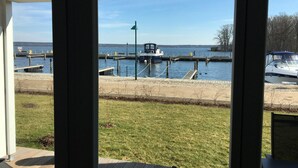 Vue depuis l’intérieur d’un bâtiment, donnant sur une marina où des bateaux sont amarrés, une zone herbeuse et un ciel dégagé.