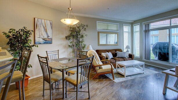 A living room with a dining table, chairs, a sofa, and a coffee table. There are windows, a plant, and a painting on the wall. - Garden level getaway at Discovery Bay (Kelowna)