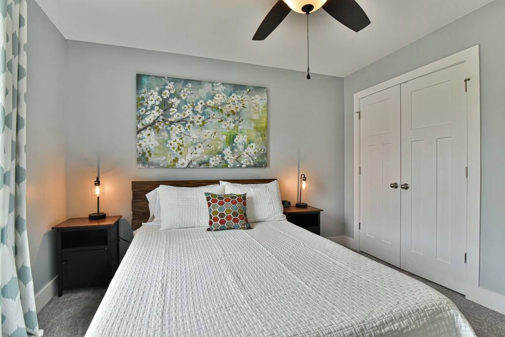 A bedroom with a large bed, two bedside tables, a ceiling fan, a painting on the wall, and a wardrobe.