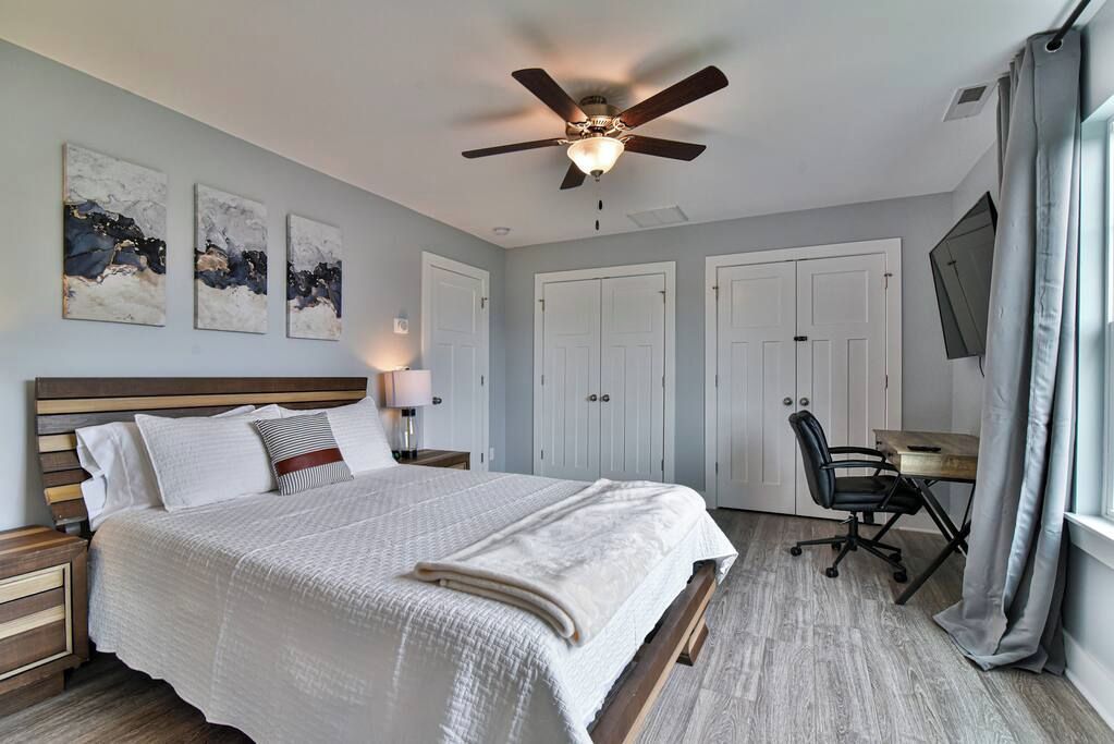 A bedroom with a bed, a desk, a chair, a ceiling fan, and artwork on the wall.