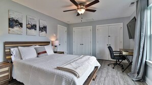 Un dormitorio con cama, escritorio, silla, ventilador de techo y obras de arte en la pared.