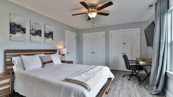 A bedroom with a bed, a desk, a chair, a ceiling fan, and artwork on the wall.