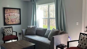 A living room with a grey sofa, two armchairs, a coffee table, a ceiling fan, and a window with curtains.