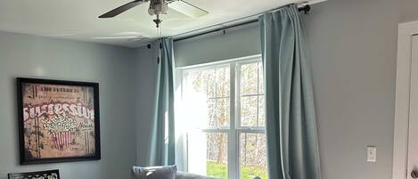 Una sala de estar con un sofá gris, dos sillones, una mesa de centro, un ventilador de techo y una ventana con cortinas.