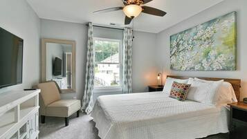 A bedroom with a bed, a ceiling fan, a window with curtains, a painting on the wall, and a TV on the left.
