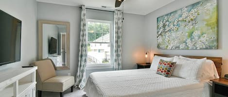 A bedroom with a bed, a ceiling fan, a painting, a window with curtains, and a TV on a stand.