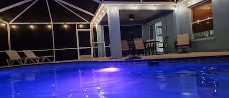 Una piscina con borde iluminado en azul, rodeada por una zona cubierta con sillas y mesas, y vista al cielo.
