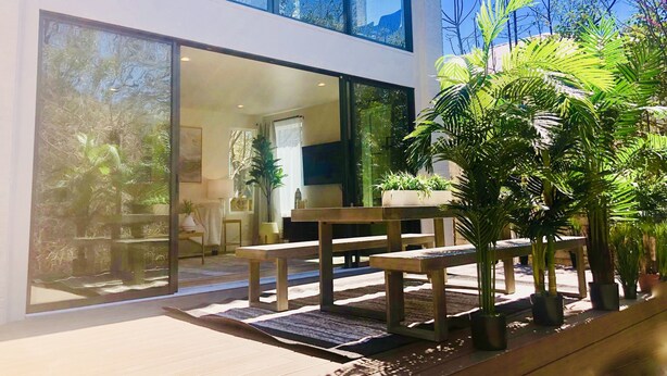 A modern two-story house with large glass windows, a patio with a wooden table and chairs, and several potted plants. - Huge Deck Overlooking Waller Creek & Lush Backyard (Austin)