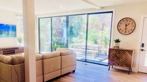 A modern living room with a large window, a sofa, a wooden cabinet, and a wall clock. - Huge Deck Overlooking Waller Creek & Lush Backyard (Austin)