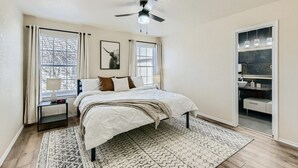 Un dormitorio con cama, mesita de noche, ventilador de techo, ventana con cortinas y vistas al exterior.