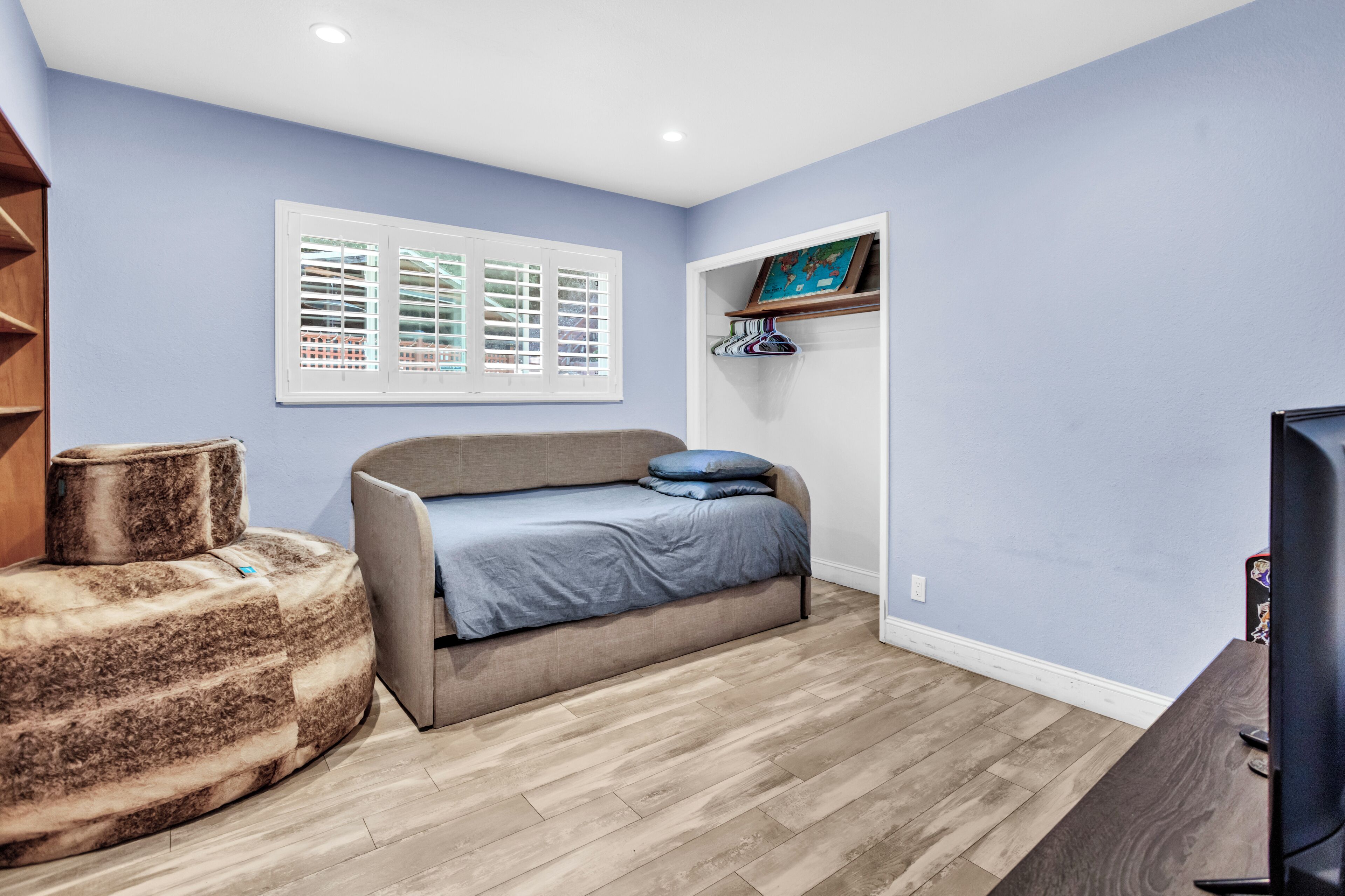 Quarto com cama, um puf, televisão, janela com persianas e prateleira com uma fotografia.
