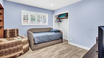 Quarto com cama, um puf, televisão, janela com persianas e prateleira com uma fotografia.
