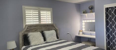 Une chambre à coucher comprenant un lit, une table de chevet, une commode, une coiffeuse avec éclairage et une fenêtre avec des stores.
