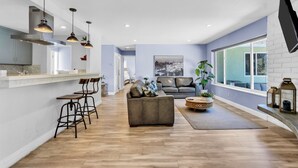 Een moderne keuken met hanglampen, een eethoek met barkrukken, een woonkamer met een bank en een groot raam, en een flatscreen-tv bevestigd aan een bakstenen muur.