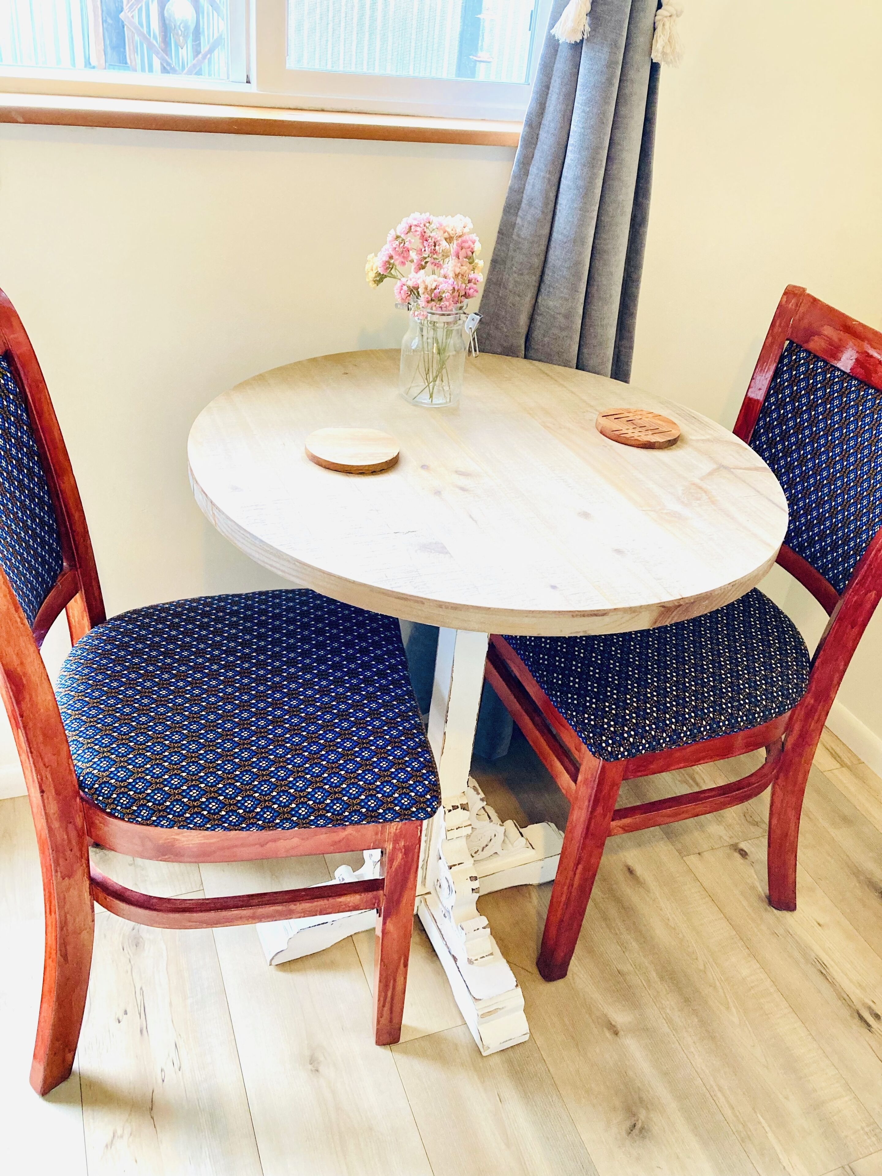 Uma mesa redonda de madeira com um vaso de flores, rodeada por quatro cadeiras com estofados estampados em azul.