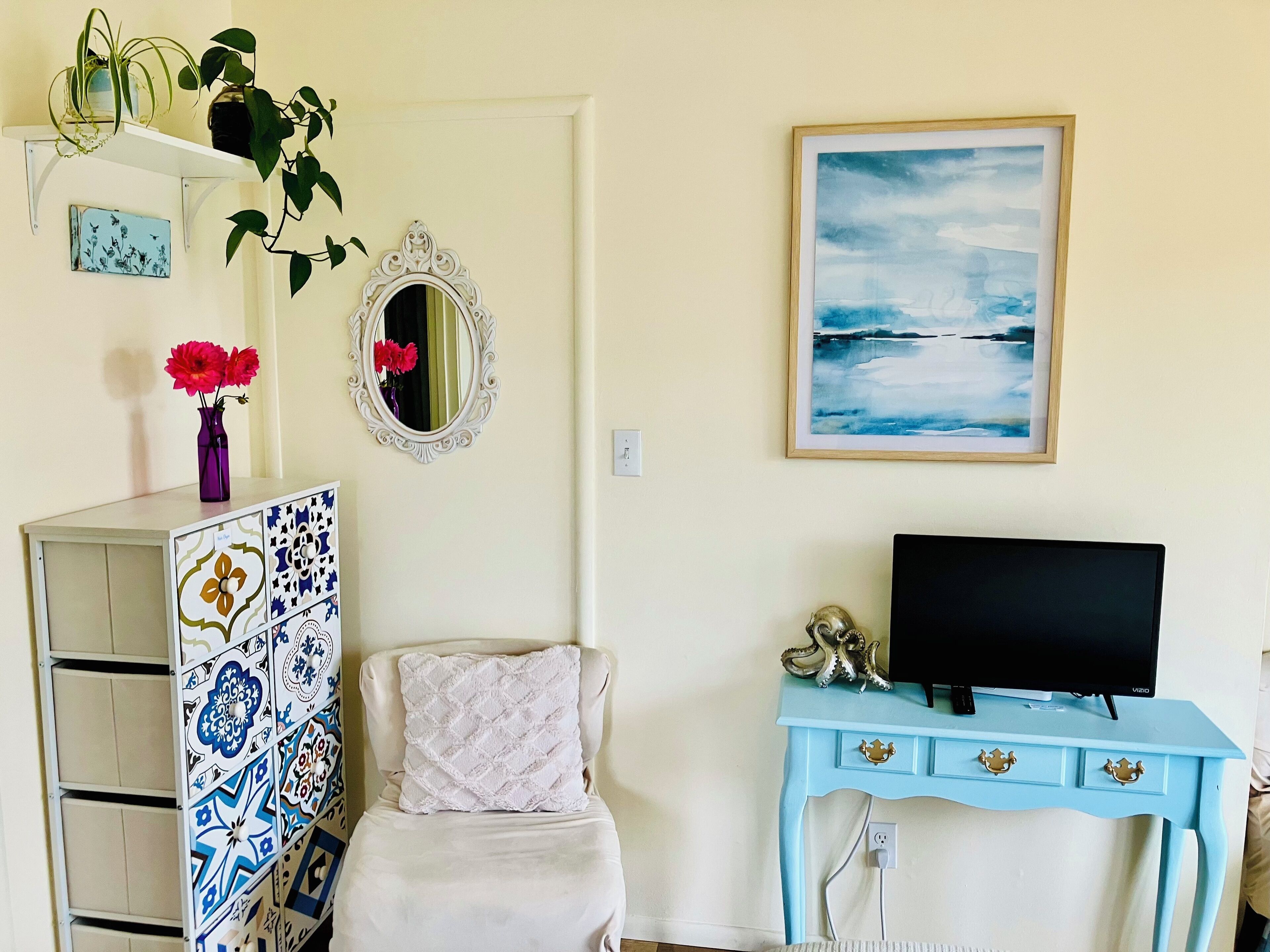 A room with a bed, a TV, a blue dresser, a mirror, and a painting of a seascape.