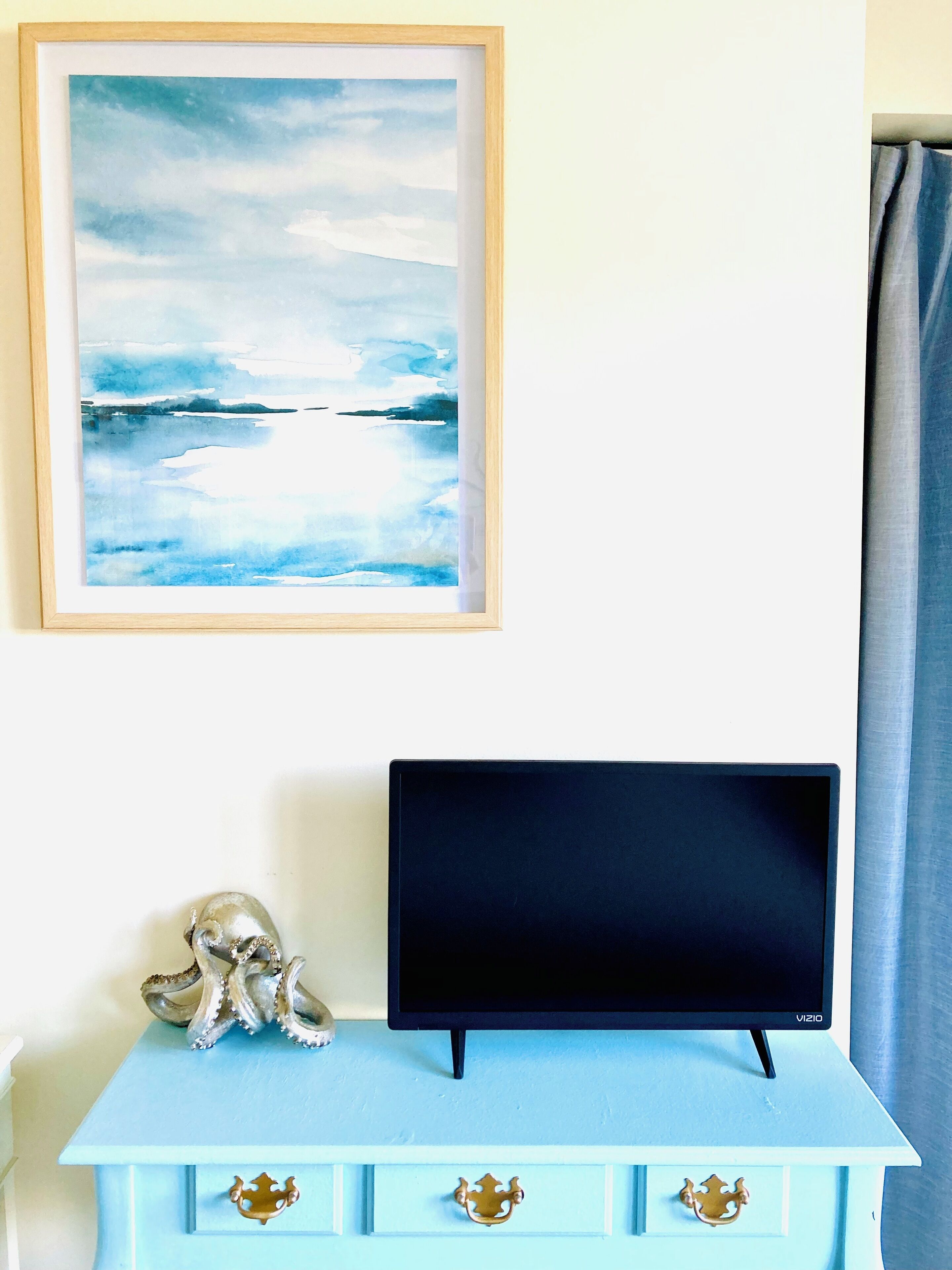 Um quarto com uma TV sobre uma cômoda azul clara, um quadro emoldurado acima e uma cortina à direita.
