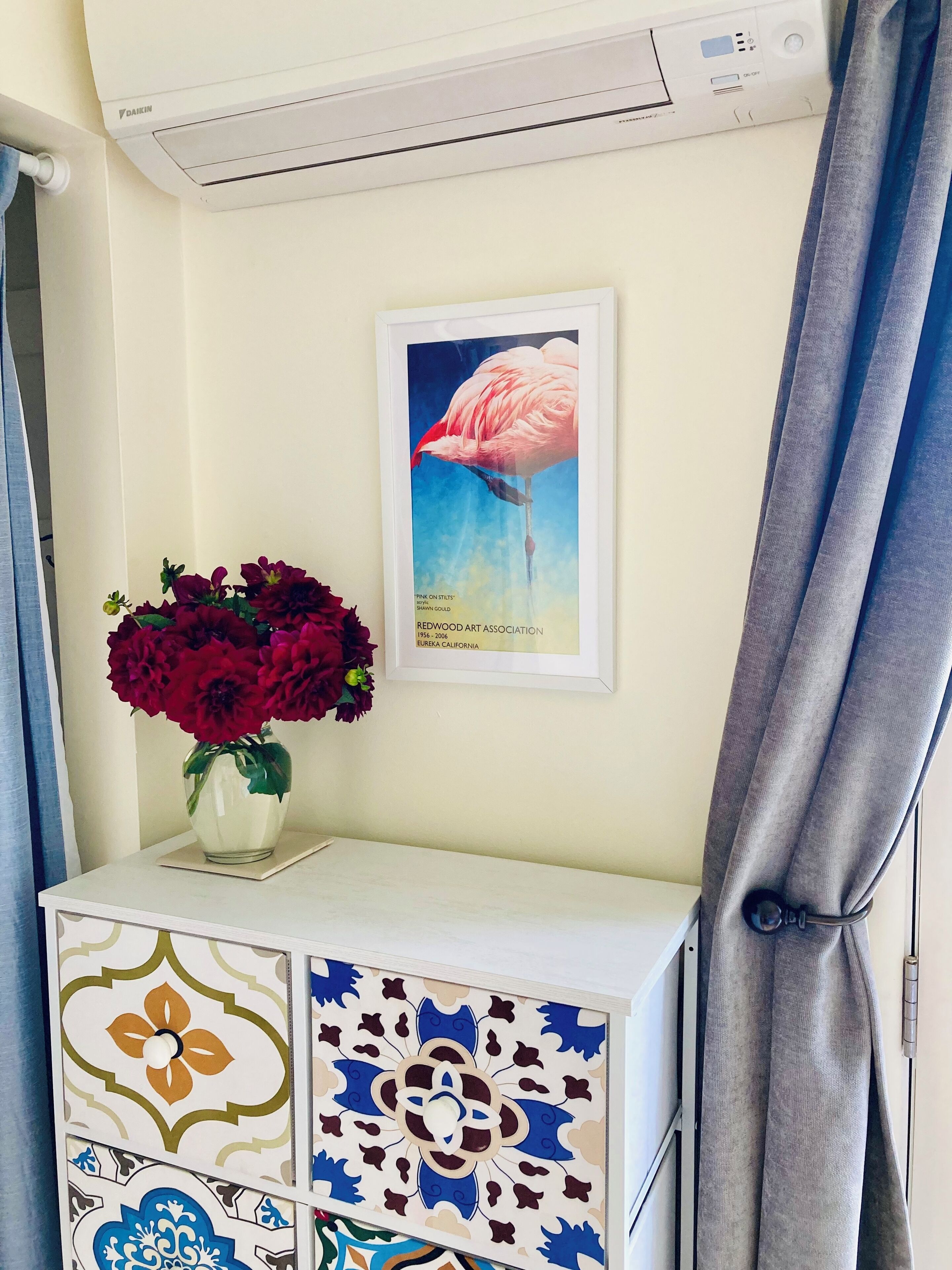 A white cabinet with a patterned tile design, a framed artwork of a flamingo, and a vase of red flowers on top.