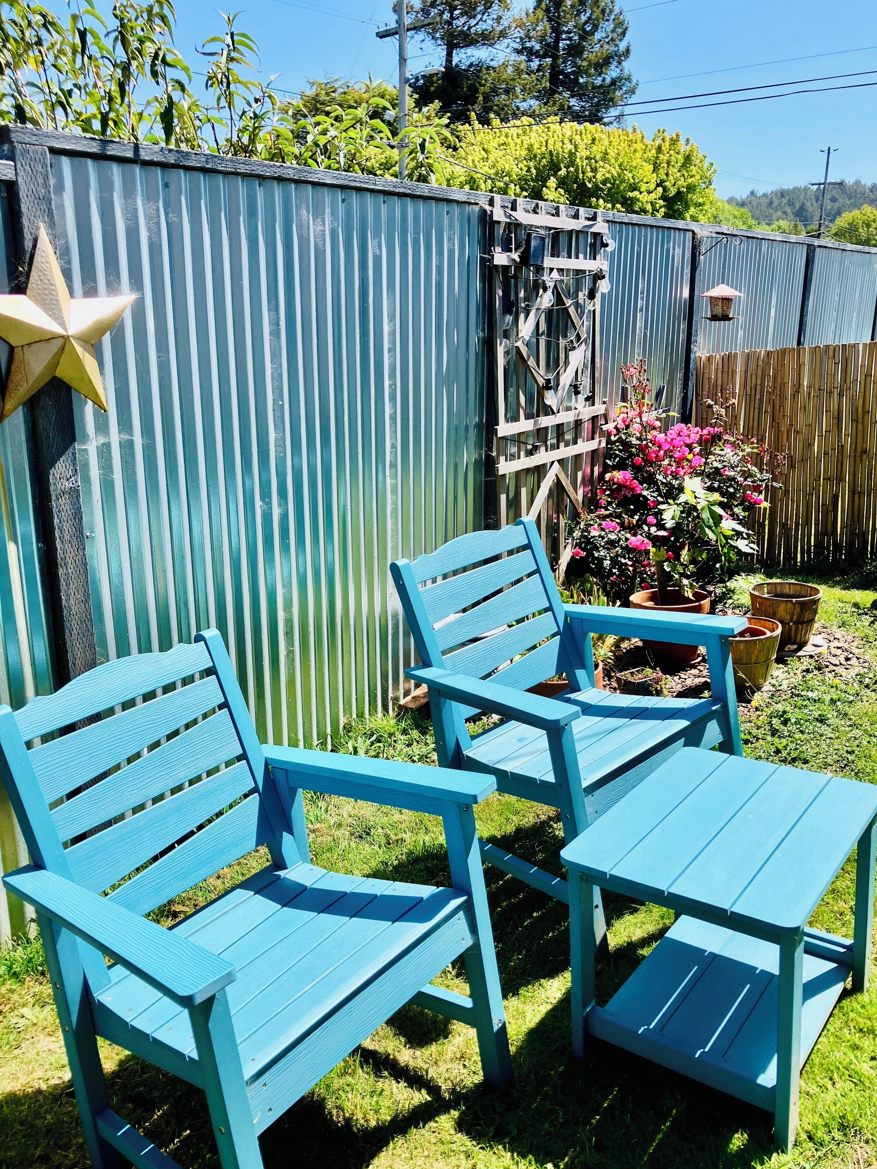 A patio area with two blue chairs and a small table, surrounded by a corrugated metal fence and a wooden fence with a climbing plant.
