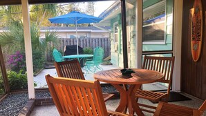 Un'area patio coperta con sedie e tavolo in legno, un ombrello blu e una decorazione a forma di tavola da surf sulla parete.