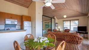 A modern kitchen with wooden cabinets, a microwave, and a stove. There is a dining area with wicker furniture and a glass table. A ceiling fan is visible, and the room has large windows. - Full 🌊 Views & 🌋Views Watch the🌅 and 🌕 rise (Koloa)