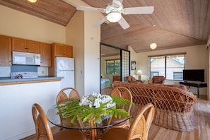 A modern kitchen with wooden cabinets, a microwave, and a stove. There is a dining area with wicker furniture and a glass table. A ceiling fan is visible, and the room has large windows.