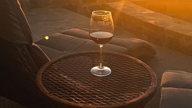 Ein Glas Rotwein auf einem Tisch mit geflochtenem Unterteil, mit Blick auf einen Sonnenuntergang über dem Meer.