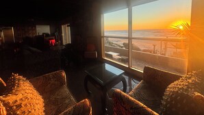 Ein Zimmer mit großem Fenster und Blick auf einen Strand bei Sonnenuntergang, mit einer Couch, einem Tisch und einem Stuhl.