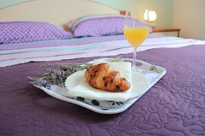 Una cama con un edredón morado, un plato con un croissant y un vaso de zumo de naranja, y un ramo de lavanda.
