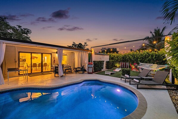 A swimming pool with a sunken seating area, a covered patio with outdoor furniture, and a well-lit garden area with string lights.